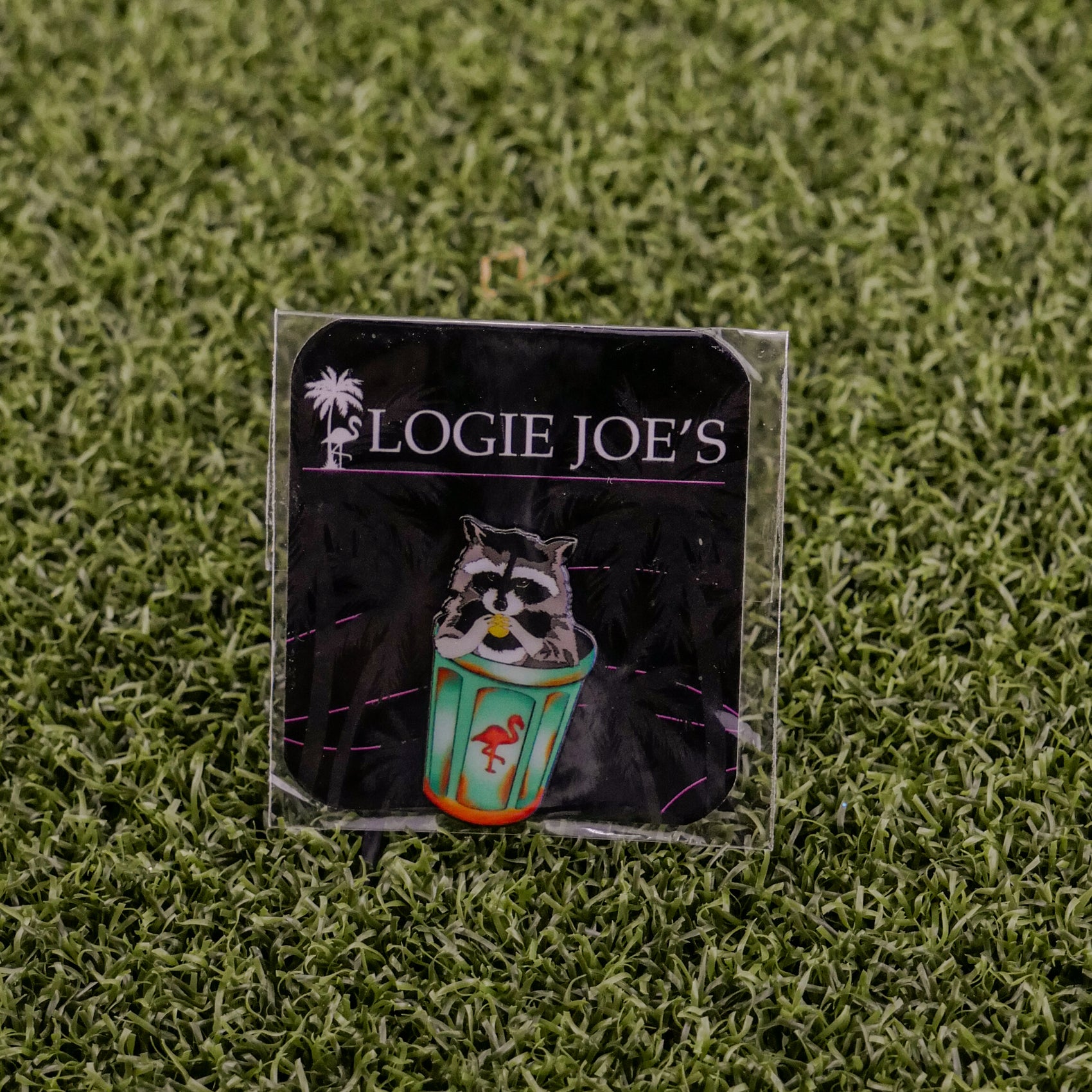 A square pin called "Trash Panda" by Logie Joe’s Golf shows a raccoon holding a colorful cup with a flamingo, set on artificial grass. The backing card features “LOGIE JOE’S” and a palm tree, perfect for premium golf accessory fans.