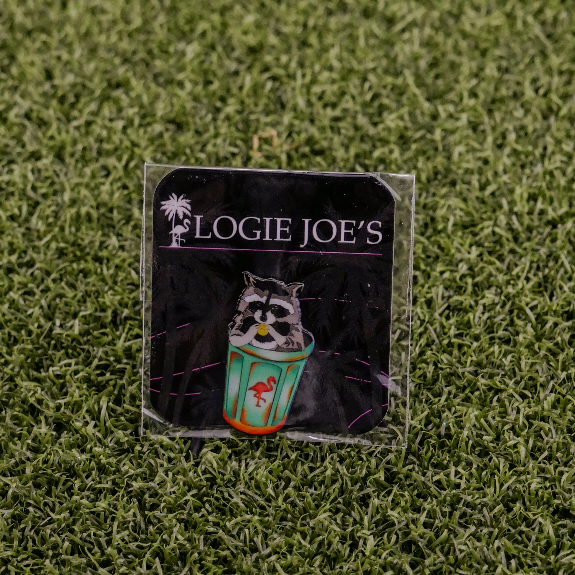 A square pin called "Trash Panda" by Logie Joe’s Golf shows a raccoon holding a colorful cup with a flamingo, set on artificial grass. The backing card features “LOGIE JOE’S” and a palm tree, perfect for premium golf accessory fans.