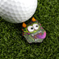 A golf ball rests on artificial grass next to Logie Joe's Golf Troll—a funny, colorful golf ball marker shaped like a cartoon monster with orange horns, big eyes, and a large open mouth with a purple tongue.