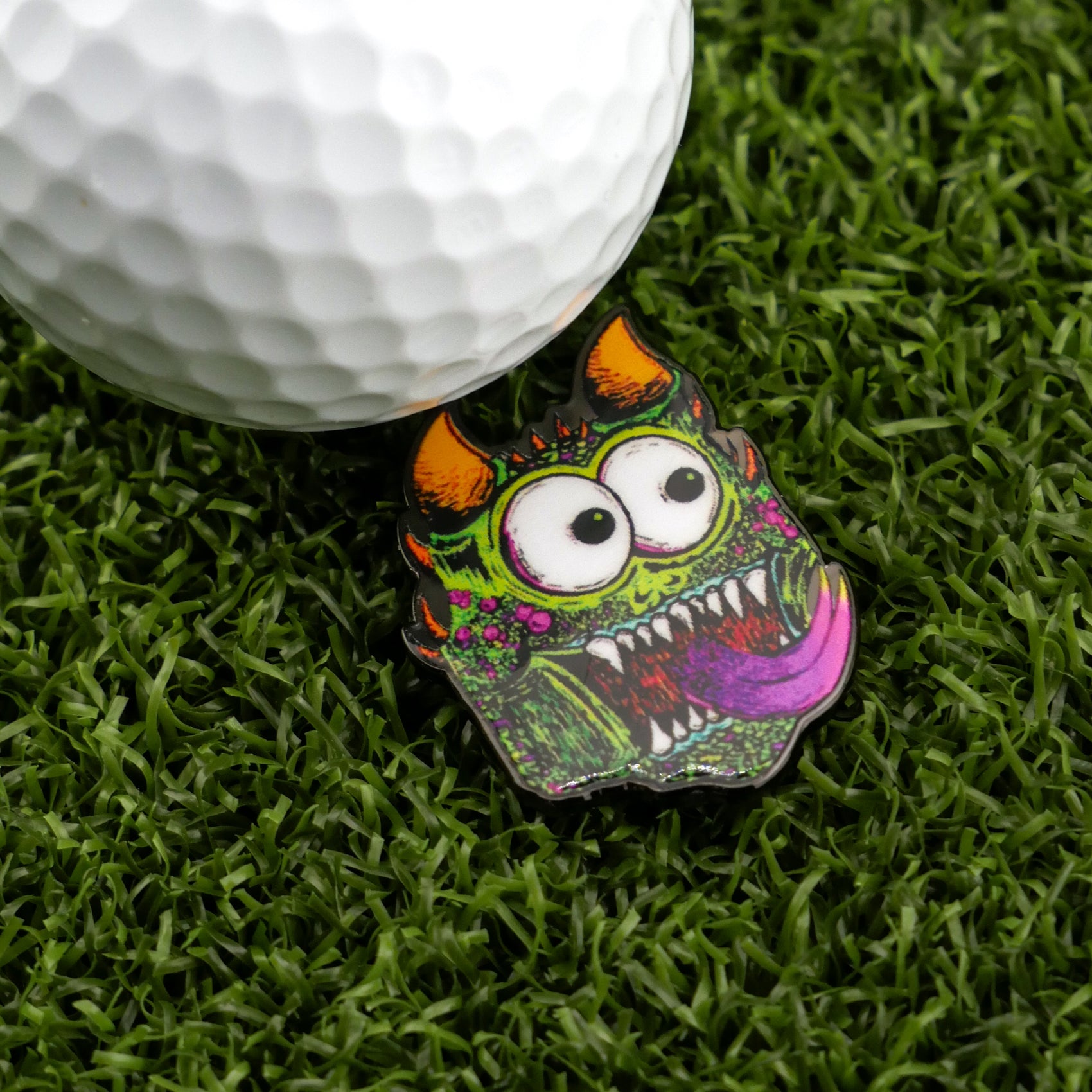 A golf ball rests on artificial grass next to Logie Joe's Golf Troll—a funny, colorful golf ball marker shaped like a cartoon monster with orange horns, big eyes, and a large open mouth with a purple tongue.