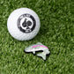 A white golf ball with the Pins and Aces logo rests on green turf beside the Trout Ball Marker, a colorful, enamel-painted fish-shaped magnetic marker by Pins and Aces.