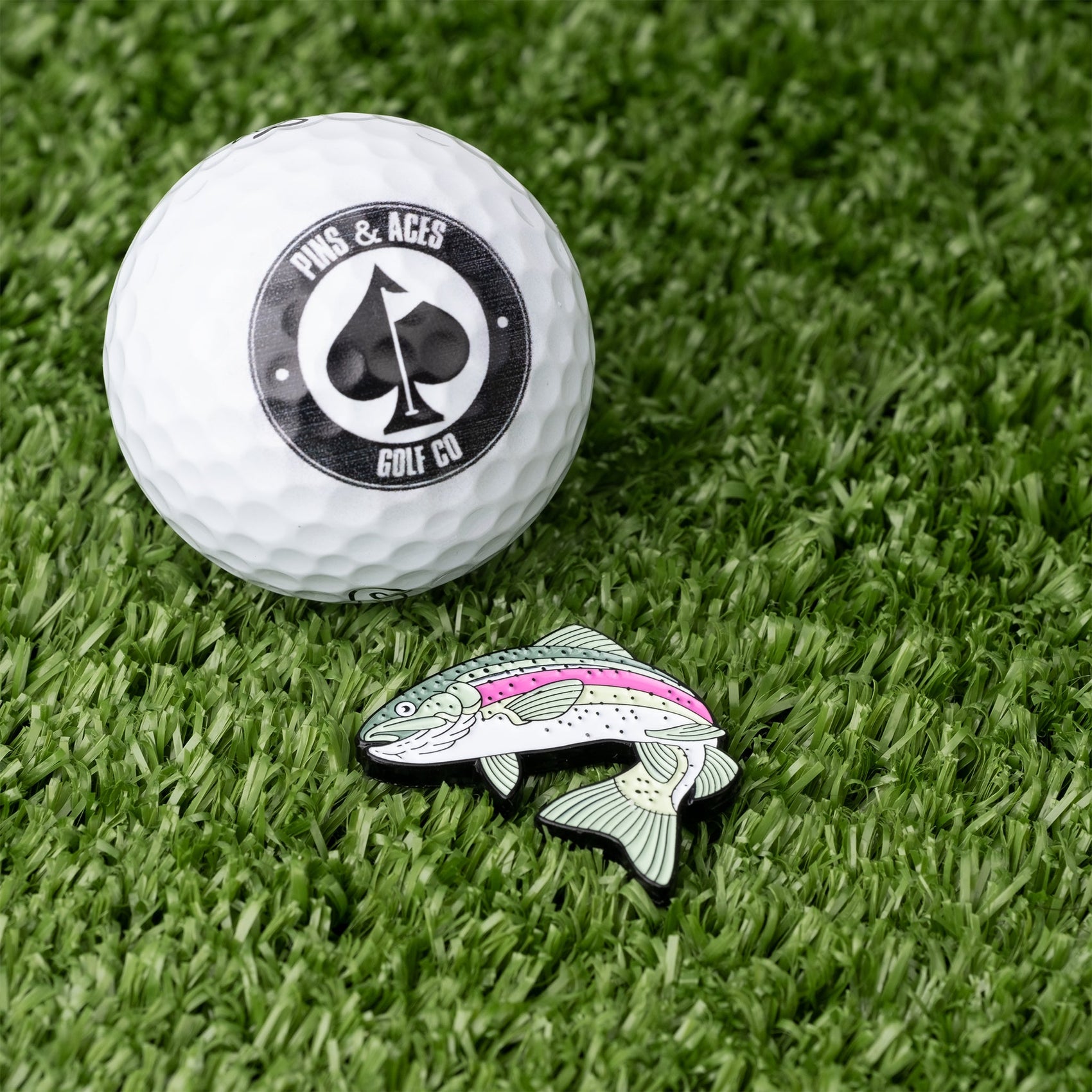 A white golf ball with the Pins and Aces logo rests on green turf beside the Trout Ball Marker, a colorful, enamel-painted fish-shaped magnetic marker by Pins and Aces.