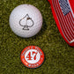 A Pins and Aces 47 Ball Marker, a limited edition red hat with a U.S. flag patch, and a golf ball with a spade symbol are displayed on green turf.