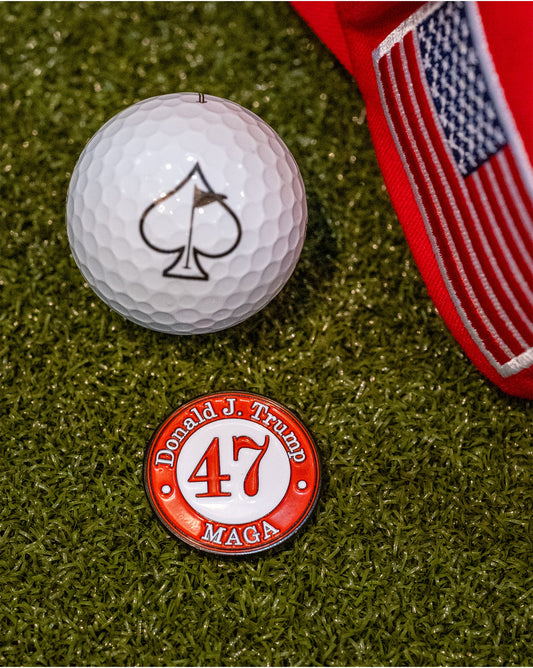 A Pins and Aces 47 Ball Marker, a limited edition red hat with a U.S. flag patch, and a golf ball with a spade symbol are displayed on green turf.