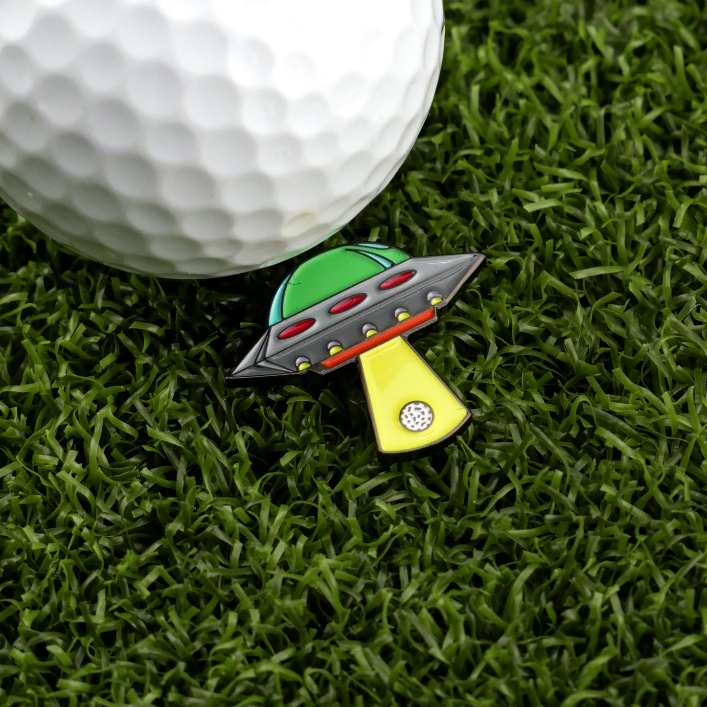 A golf ball rests on green grass beside Logie Joe's Golf UFO—a colorful, premium, UFO-shaped ball marker with a yellow beam pointing toward the ball.