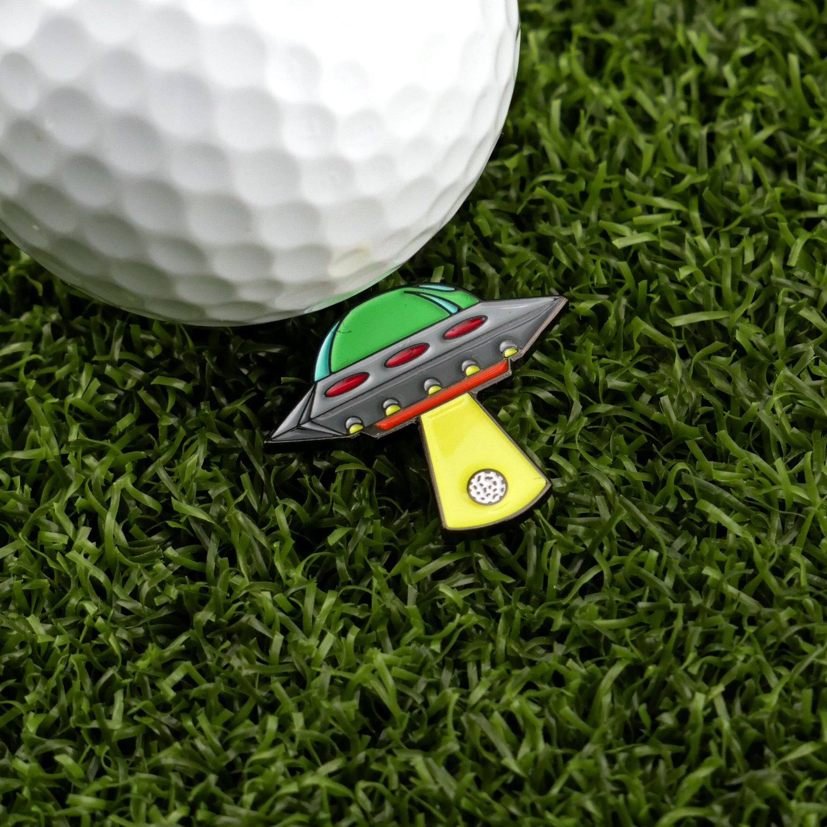 A golf ball rests on green grass beside Logie Joe's Golf UFO—a colorful, premium, UFO-shaped ball marker with a yellow beam pointing toward the ball.