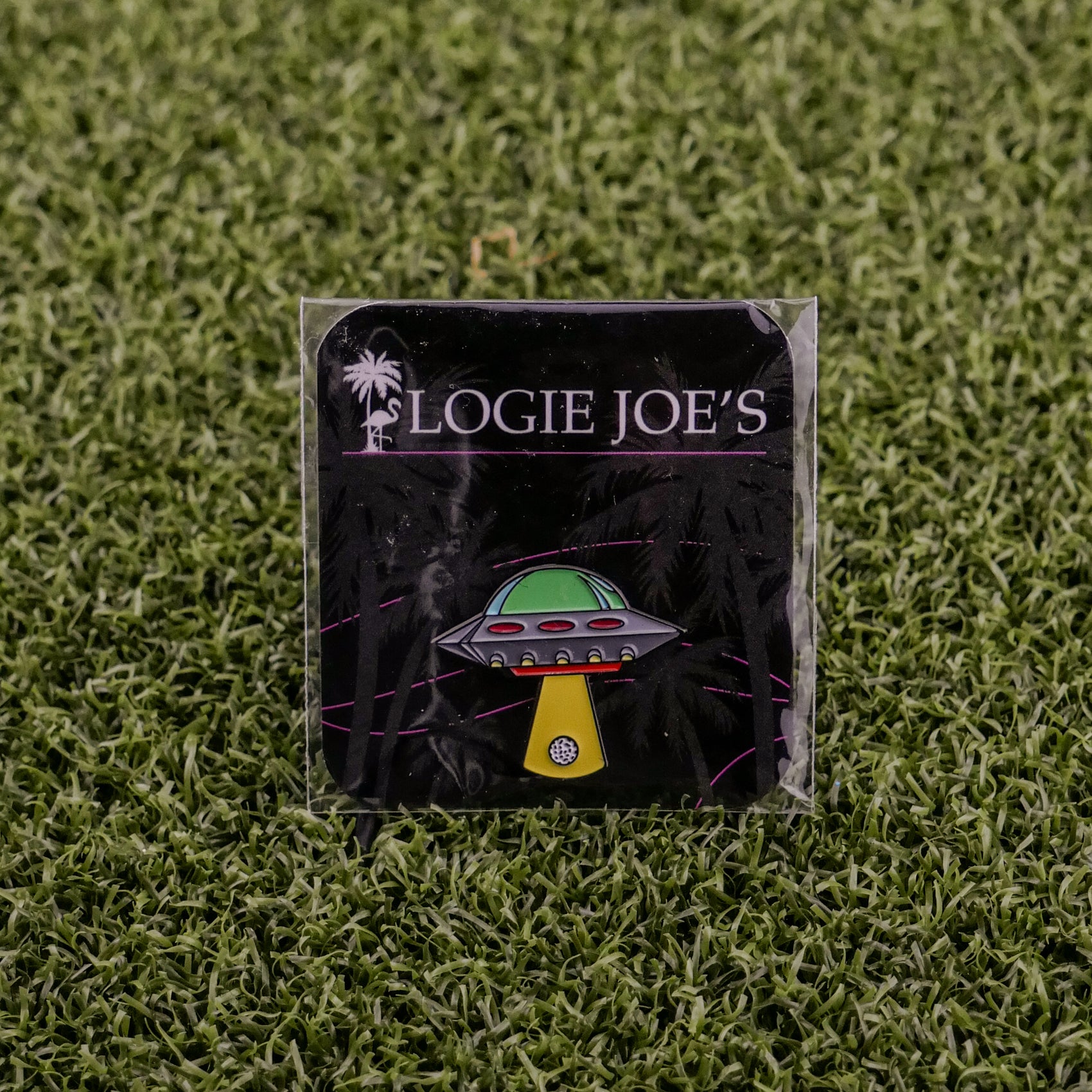 The UFO pin from Logie Joe's Golf features a green dome and yellow beam, perfect as an artistic golf accessory or unique ball marker. Displayed on a branded card, it's shown on green artificial grass for added style.