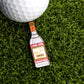 The Russian Vodka Ball Marker by Logie Joe's Golf, shaped like a vodka bottle, sits on green artificial grass beside a white golf ball—an ideal novelty golf gift and unique accessory for golfers.