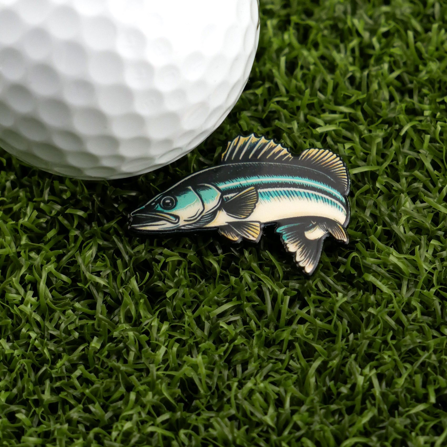 A close-up of a golf ball on artificial grass beside Logie Joe's Golf "Walleye" pin—a blue, green, and white fish-shaped enamel marker for anyone who loves funny golf accessories.