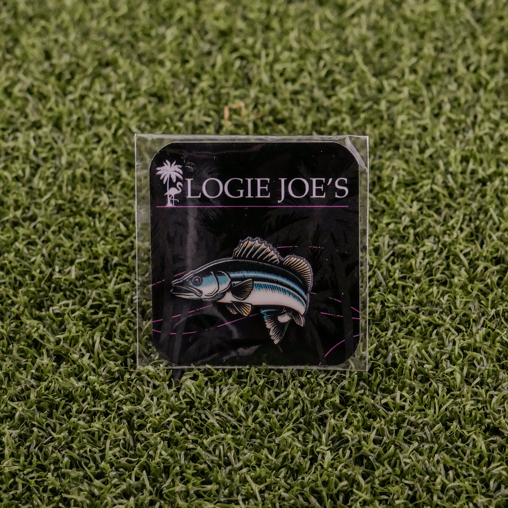 A square sticker featuring a walleye fish illustration and Logie Joe's Golf text on a dark background, set on green turf—ideal for golf enthusiasts seeking fun accessories or premium ball marker inspiration.
