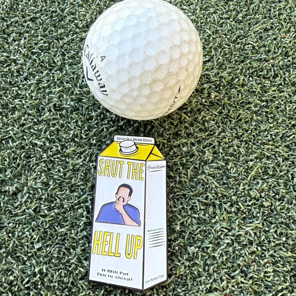 A Warm Glass Ball Marker by Grunk Dolfer sits on artificial turf beside a novelty carton labeled SHUT THE HELL UP with a cartoon shushing—bringing humor to golf games and making a unique gift that says, "It will put you to sleep!.