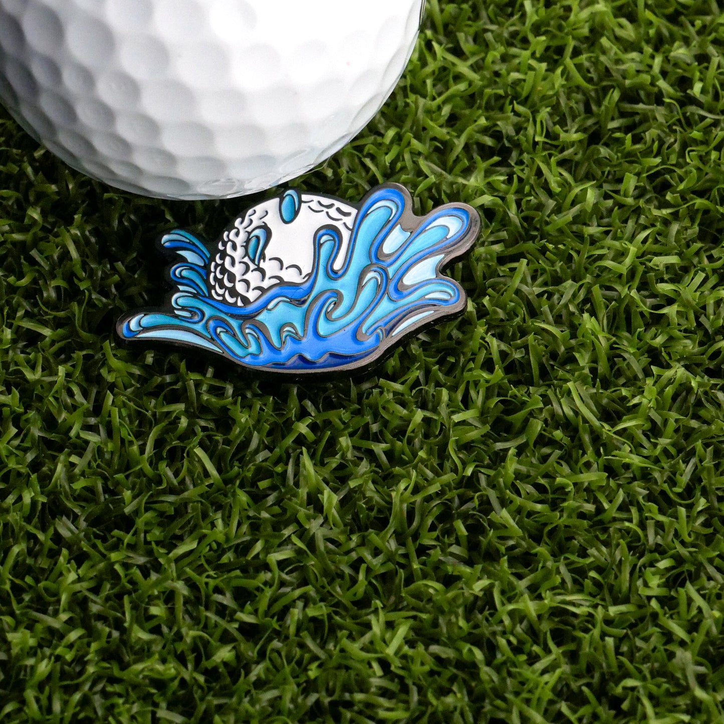 A close-up of a golf ball on artificial grass alongside the Ballmarkers.com Water Ball Ball Marker, a playful accessory shaped like a splash—perfect for golfers with a sense of humor about water hazards.