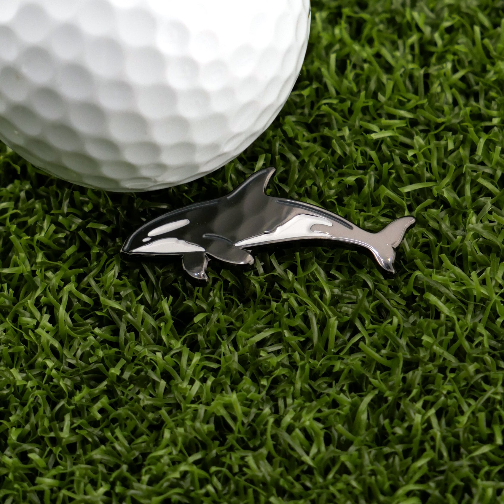 A Killer Whale-shaped golf ball marker by Logie Joe's Golf sits on green artificial grass beside a white golf ball, bringing a playful touch to your golf accessories.