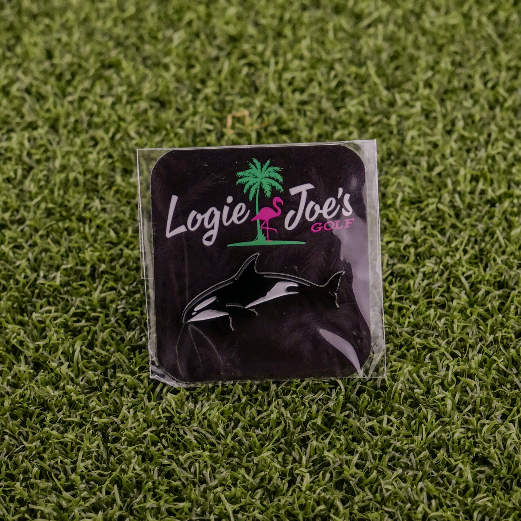 A black square pin from Logie Joe's Golf, named Killer Whale, features playful animal designs and is a humorous golf accessory—ideal as a ball marker—shown on green artificial grass.
