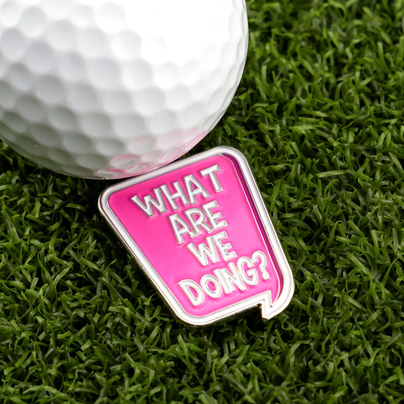 A golf ball sits on green grass beside Logie Joe's Golf "What are we doing?" Speech Bubble marker, a pink enamel accessory shaped like a speech bubble with the phrase in white letters.