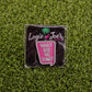 A premium Logie Joe's Golf "What are we doing?" Speech Bubble pin features a flamingo and palm tree, styled as a chic golf ball marker, shown on artificial grass alongside a pink cup engraved with the same playful phrase.
