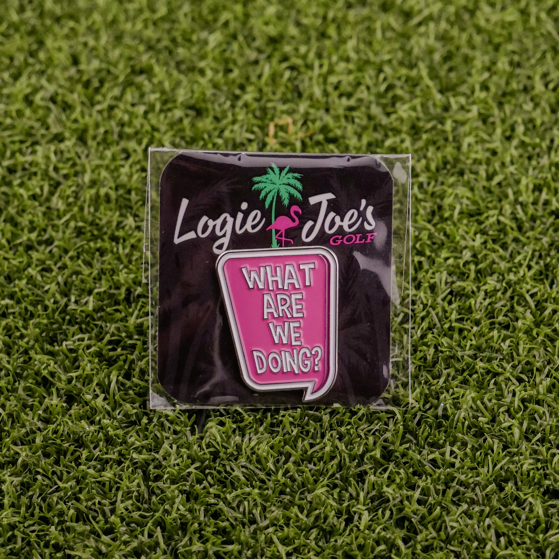 A premium Logie Joe's Golf "What are we doing?" Speech Bubble pin features a flamingo and palm tree, styled as a chic golf ball marker, shown on artificial grass alongside a pink cup engraved with the same playful phrase.