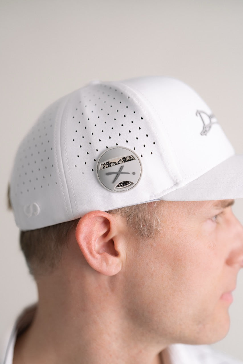 A man in profile wears Doubloon's The Omen Classic Antique, Diamond & Silver Edition—a white perforated baseball cap with a sterling silver, engraved golf ball marker—set against a plain background.