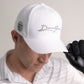 A person in a white "Darkfour" cap, black golf glove, and white collared shirt tilts their cap down, partially hiding their face, while holding Doubloon’s The Omen Classic Antique, Diamond & Silver Edition sterling silver golf ball marker.