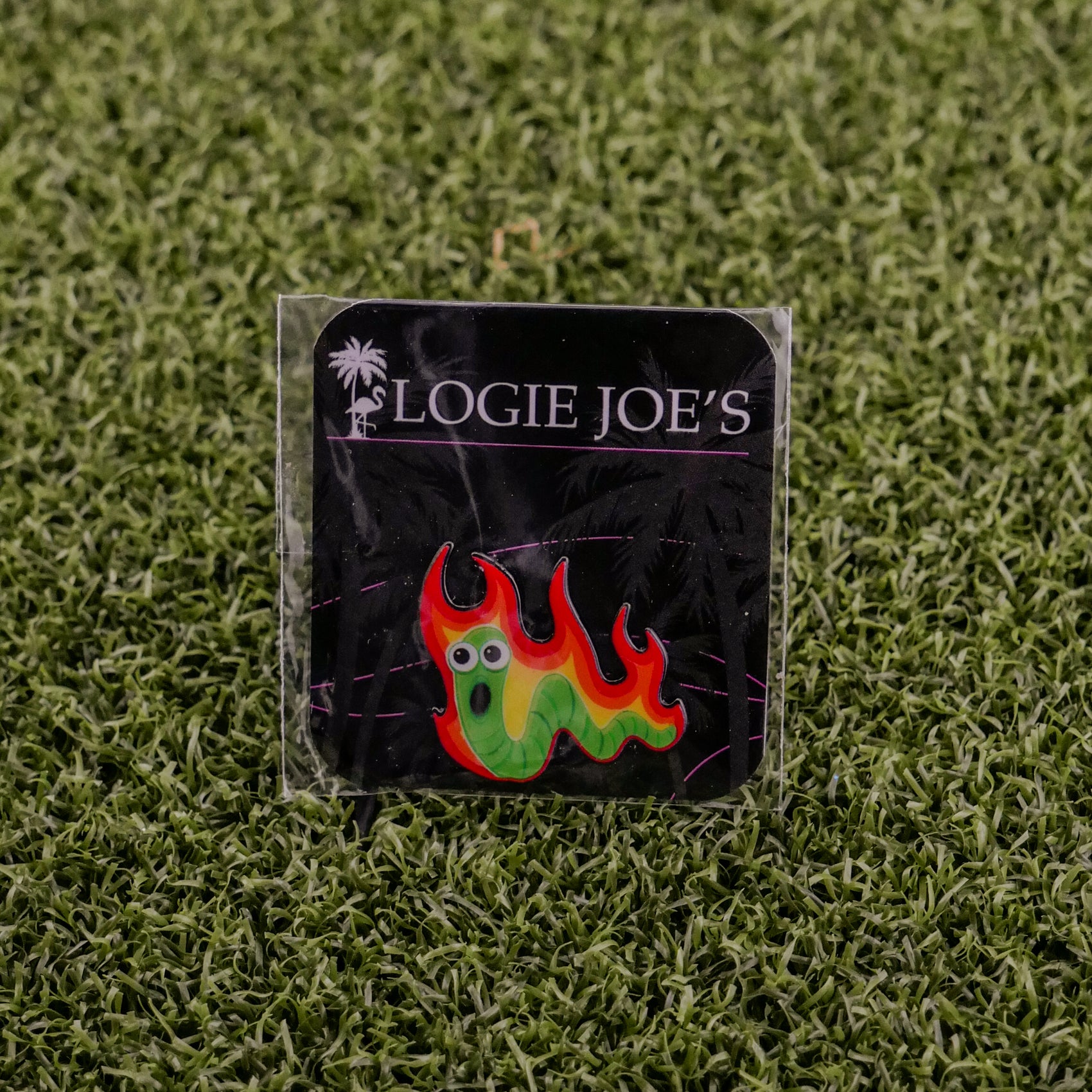 The Worm Burner by Logie Joe's Golf: a colorful enamel golf ball marker featuring a green worm with red and yellow flames and cartoon eyes, attached to a black brand card and displayed on green artificial grass—perfect for unique golf accessory fans.