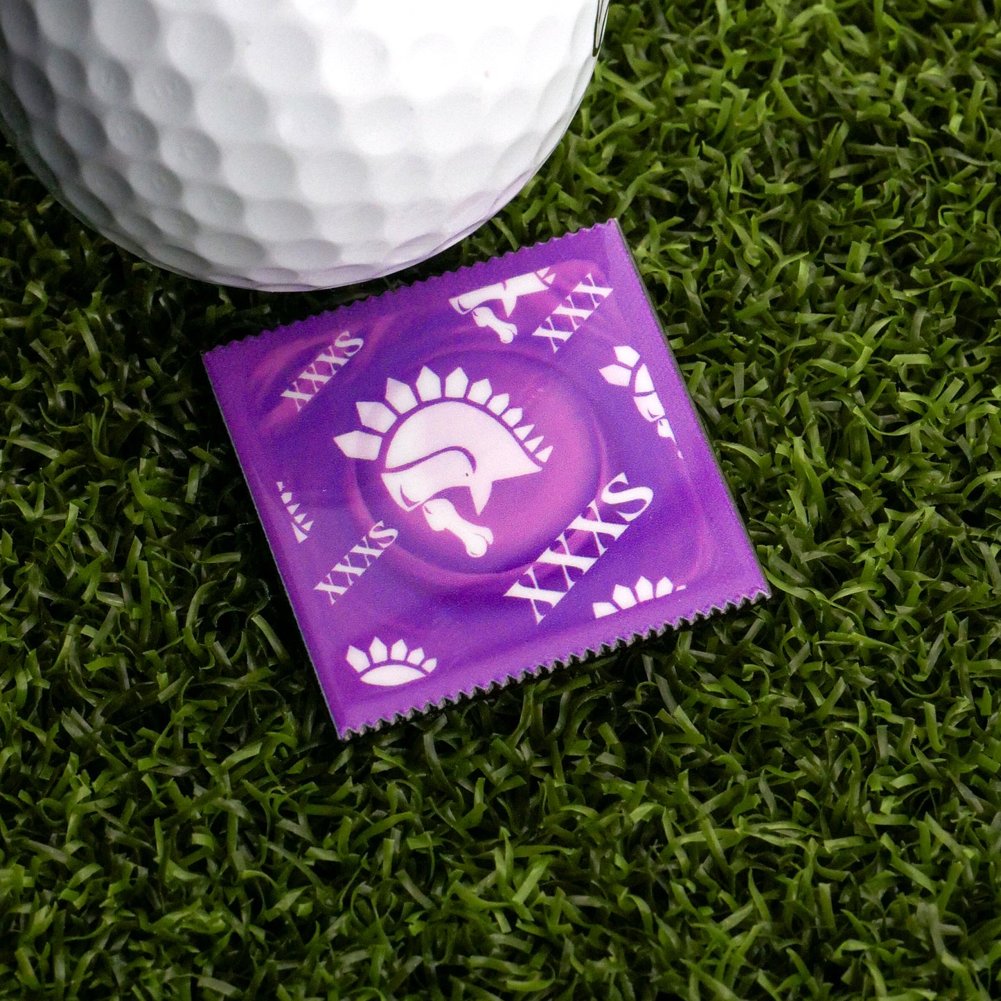 A purple XXXS Condom Ball Marker from Ballmarkers.com with white designs rests on green artificial grass next to a golf ball—perfect for golfers seeking novelty, not those aiming for Magnum XL or upset by lip outs.