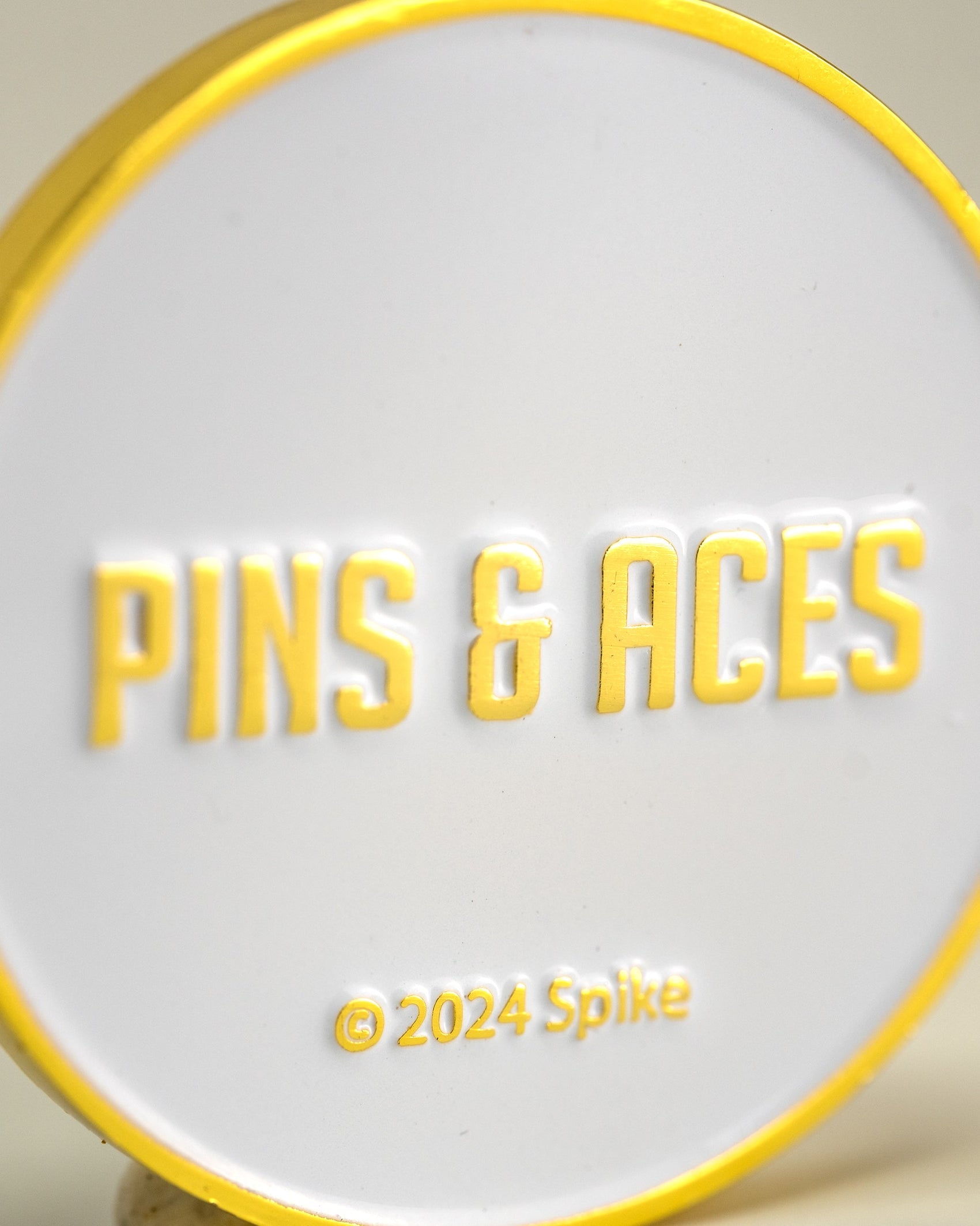 The Yellowstone Ball Marker by Pins and Aces features a round white and yellow design with raised yellow "PINS & ACES" text and © 2024 Spike—an ideal golf accessory for fans of unique collaborations.