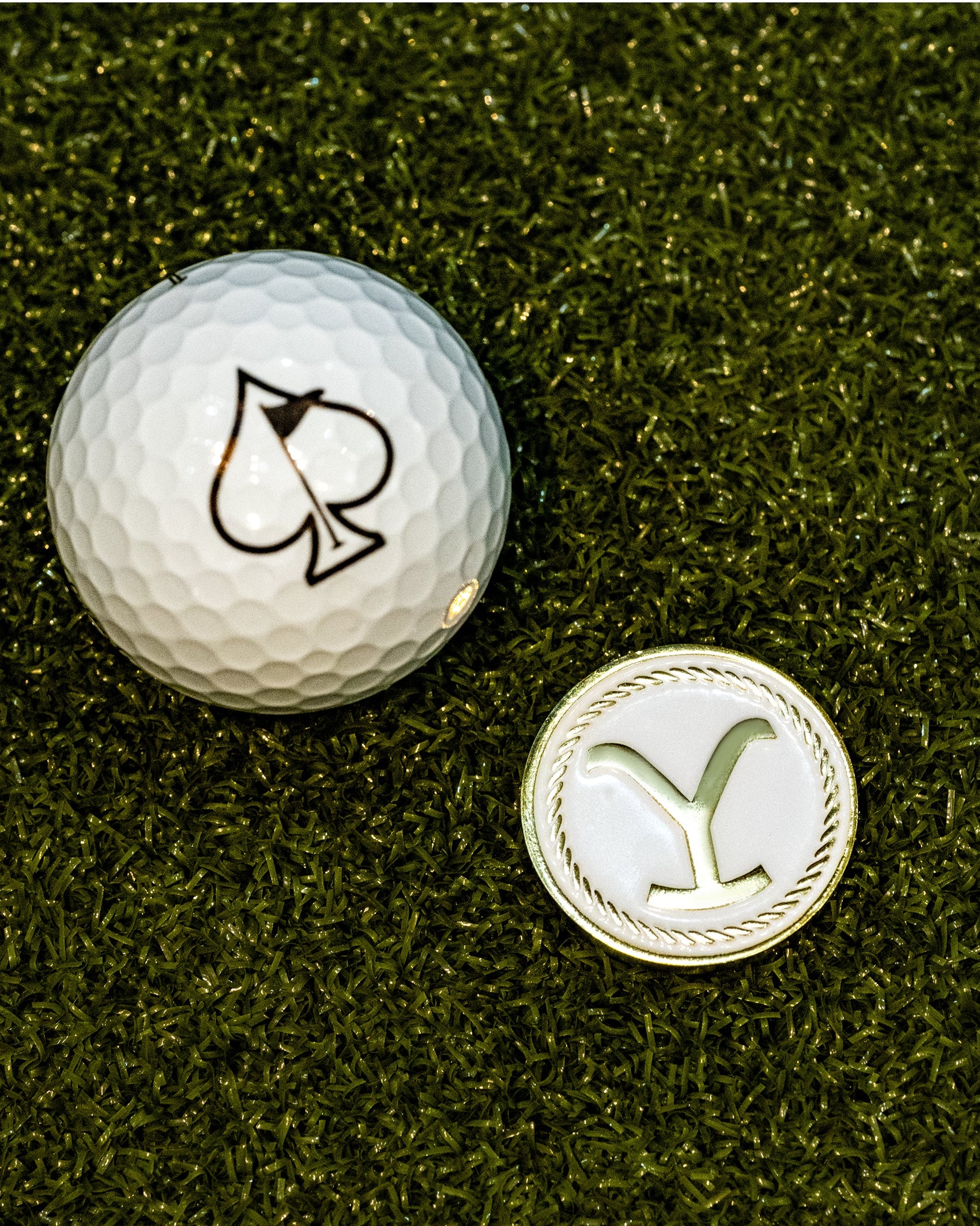A golf ball with a black spade sits next to the Yellowstone Ball Marker—with its Y-shaped design—by Pins and Aces, both on green turf. This unique golf accessory set is a highlight of the collection.