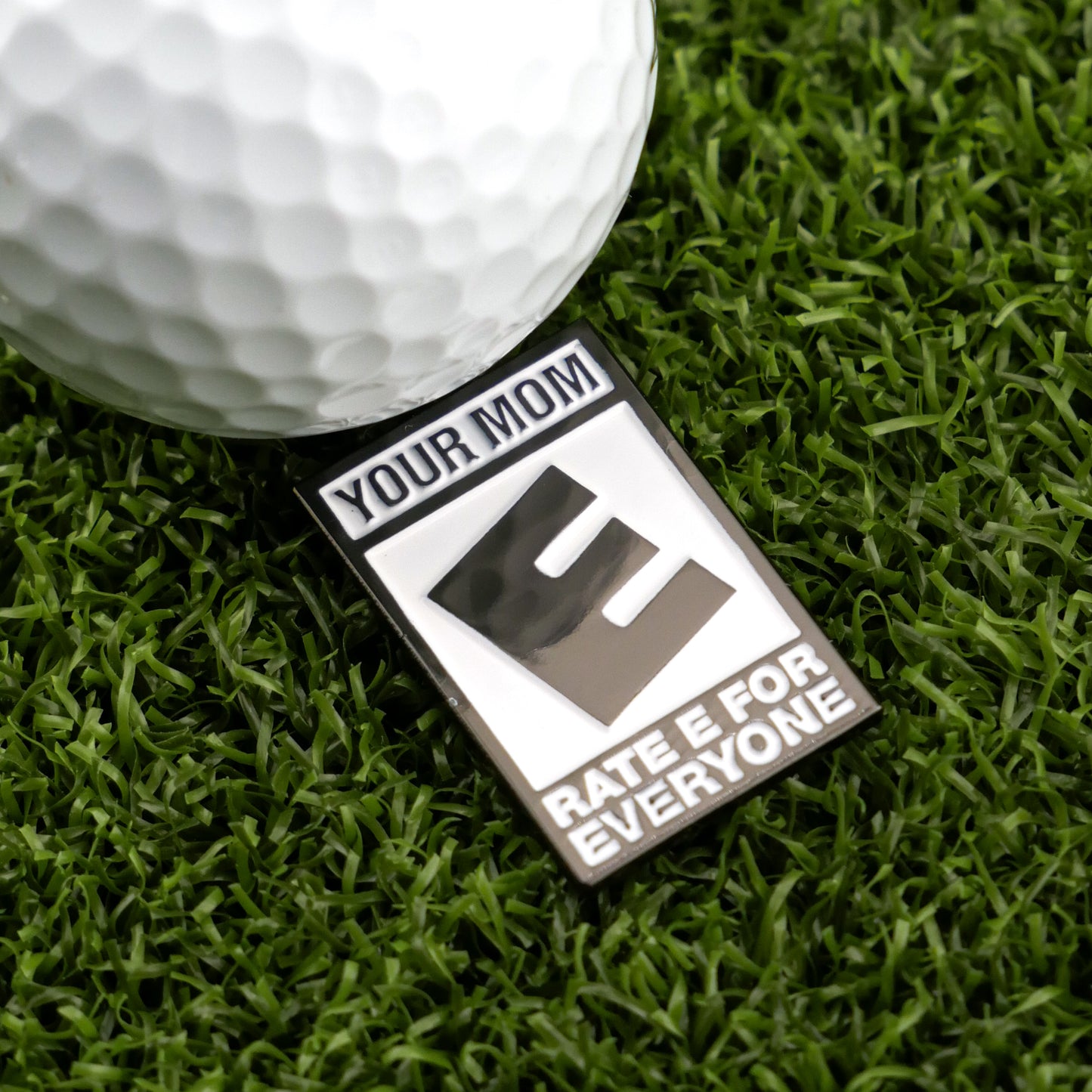 A close-up of Logie Joe's Golf "Your Mom (E For Everyone)" metallic badge and golf ball on artificial grass—ideal for fans of unique golf ball markers—engraved with YOUR MOM, a bold E, and RATE E FOR EVERYONE.