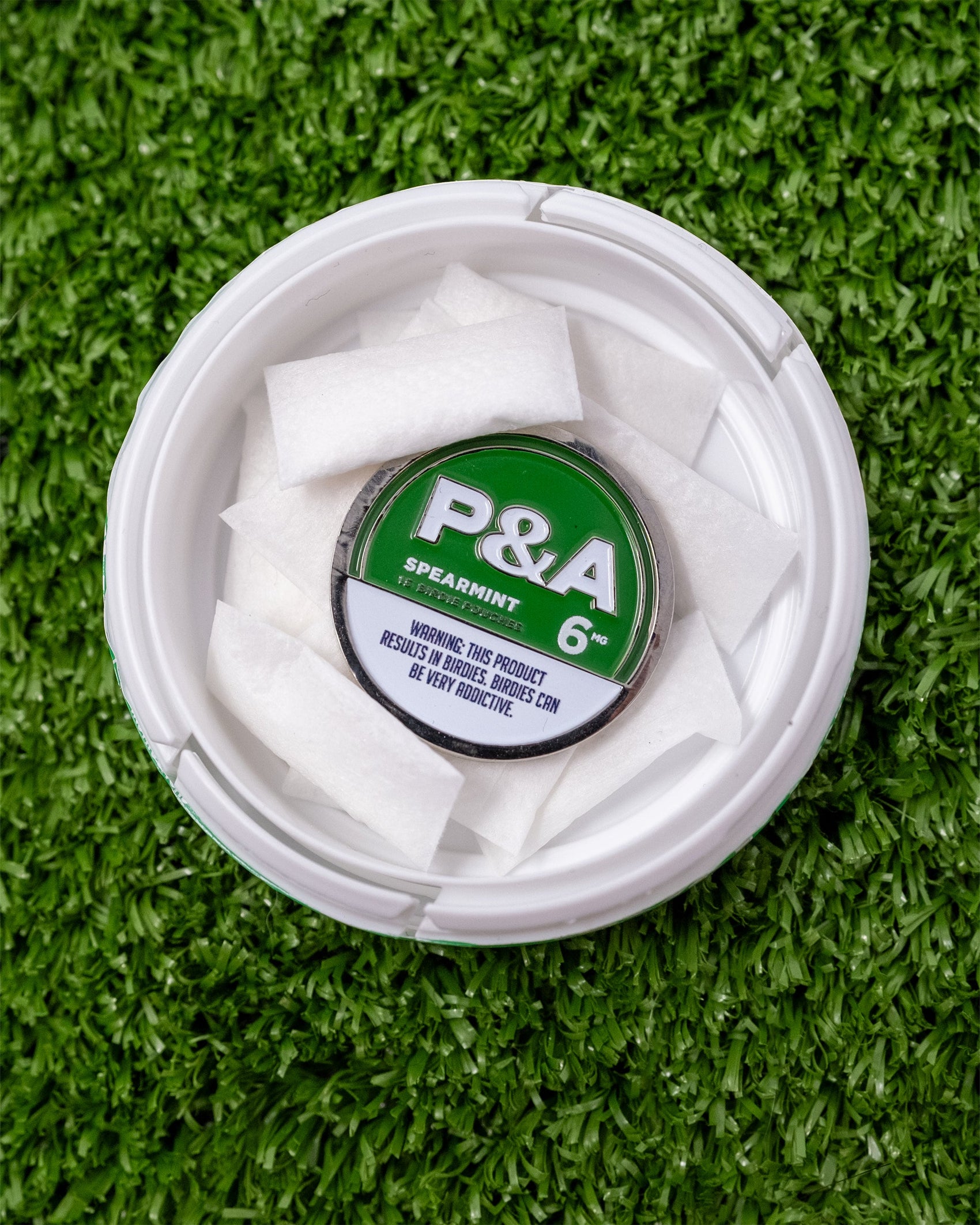 An open 6 Milly - Spearmint Ball Marker by Pins and Aces, with white rectangular pouches inside, rests on artificial green grass next to a magnetic ball marker. The product warning text appears on the lid.