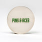 The 6 Milly - Wintergreen Ball Marker by Pins and Aces is a round, silver-colored enamel marker with "PINS & ACES" in green on the front, shown on a plain white background.