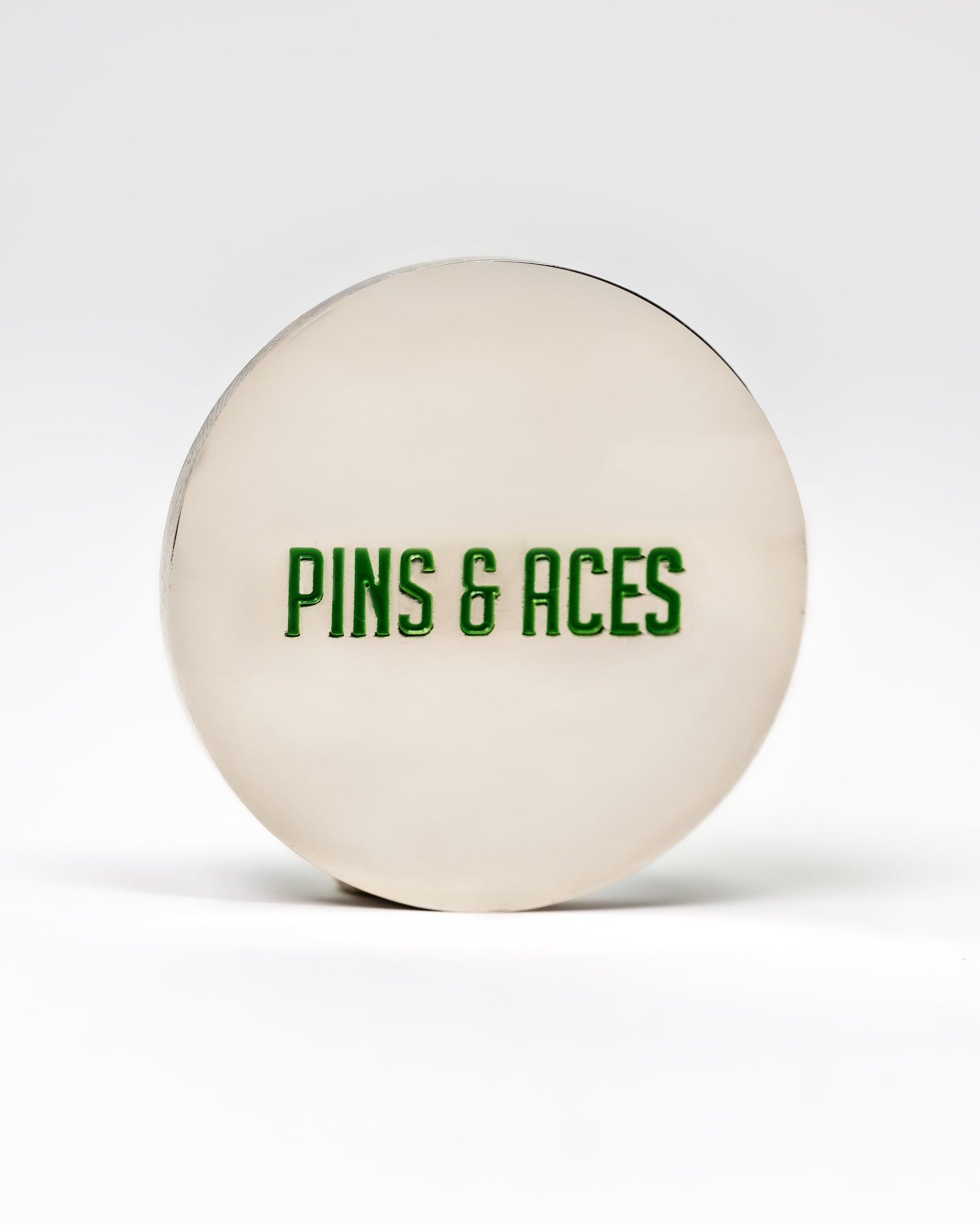The 6 Milly - Wintergreen Ball Marker by Pins and Aces is a round, silver-colored enamel marker with "PINS & ACES" in green on the front, shown on a plain white background.