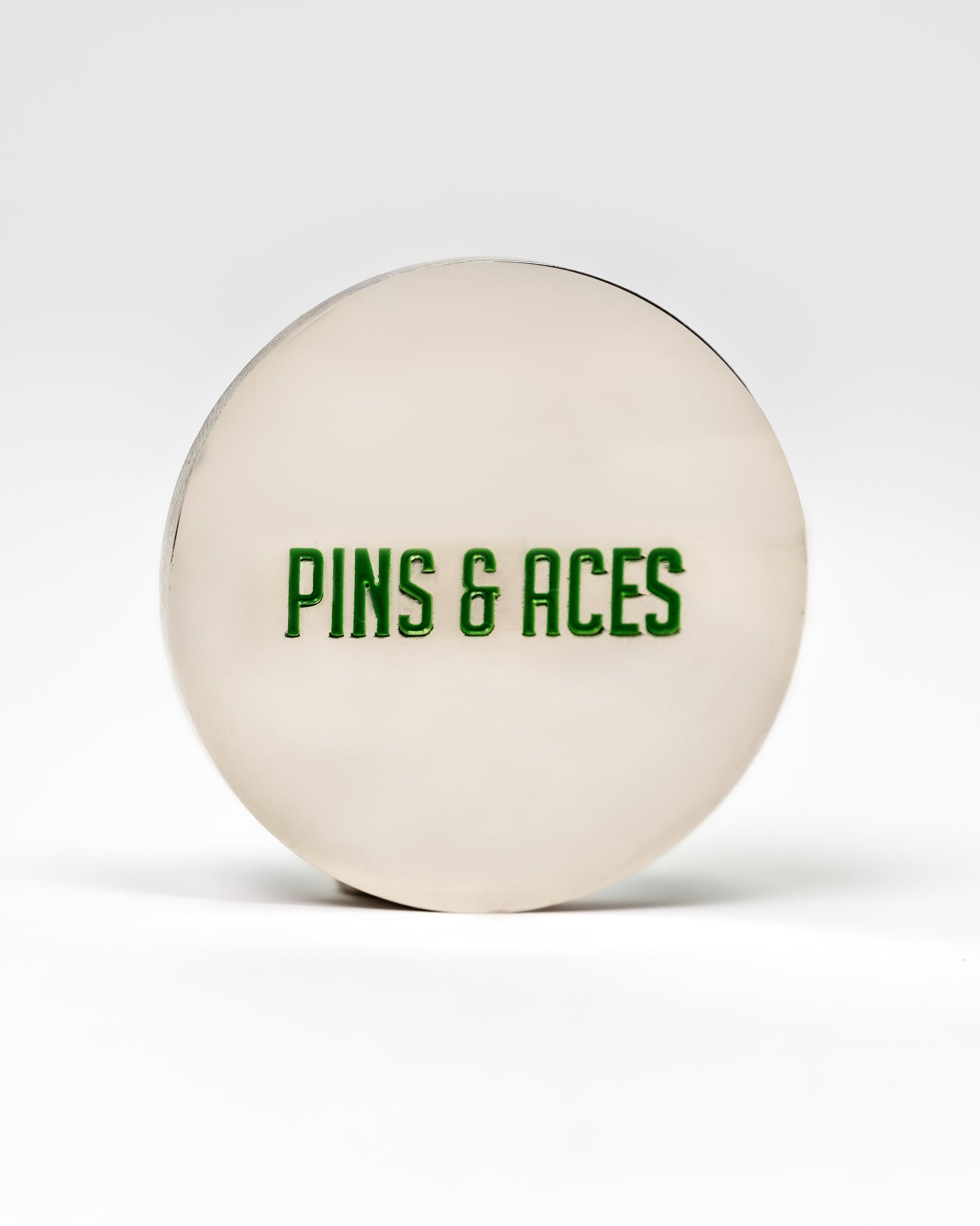 The 6 Milly - Wintergreen Ball Marker by Pins and Aces is a round, silver-colored enamel marker with "PINS & ACES" in green on the front, shown on a plain white background.