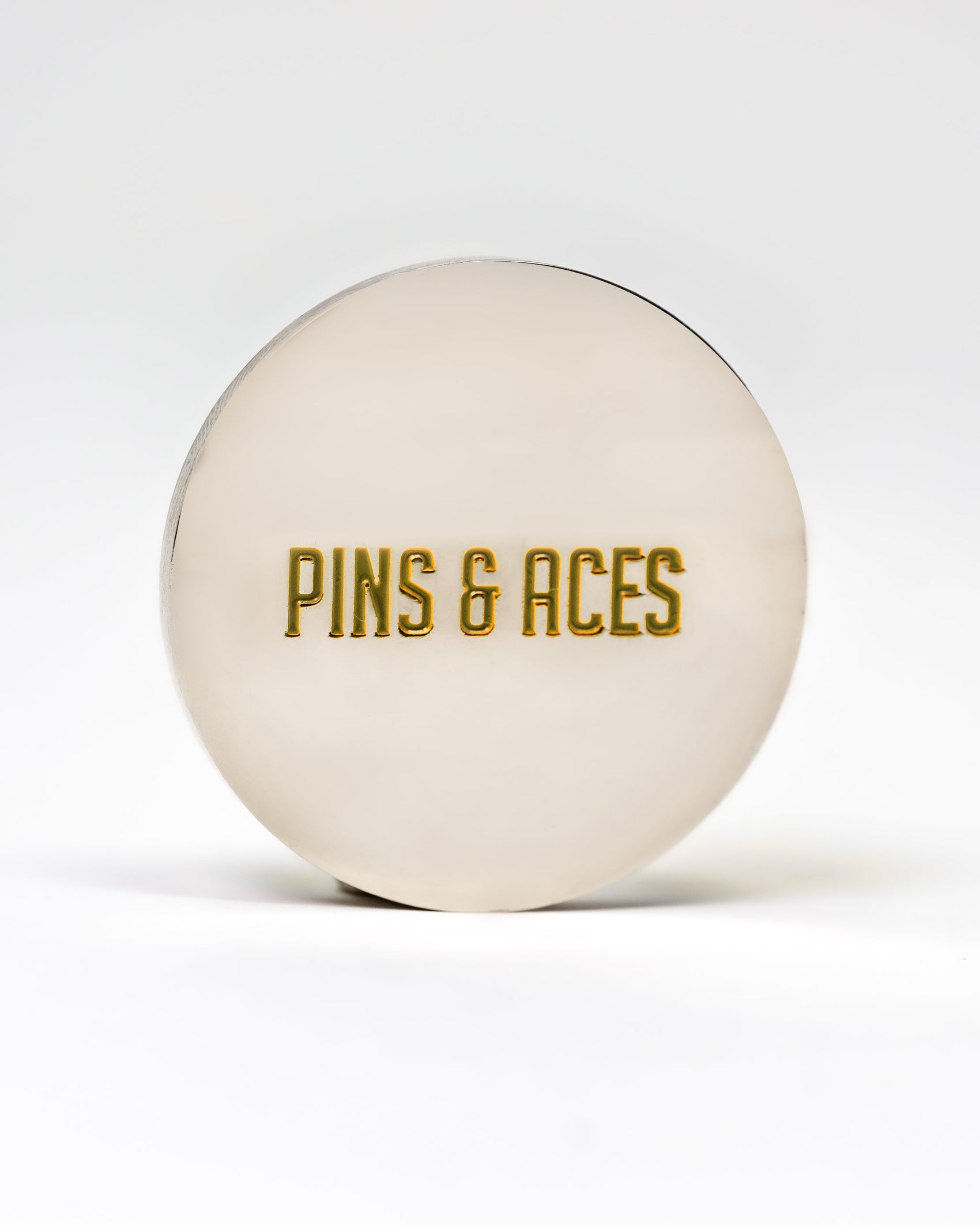 A round, metallic 6 Milly - Cinnamon Ball Marker by Pins and Aces features gold engraved branding and a smooth enamel finish, shown against a plain white background.