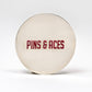 The 6 Milly - Smooth Ball Marker by Pins and Aces is a round, metallic marker with bold red "PINS & ACES" lettering on the front, finished with enamel paint and displayed on a plain white background.