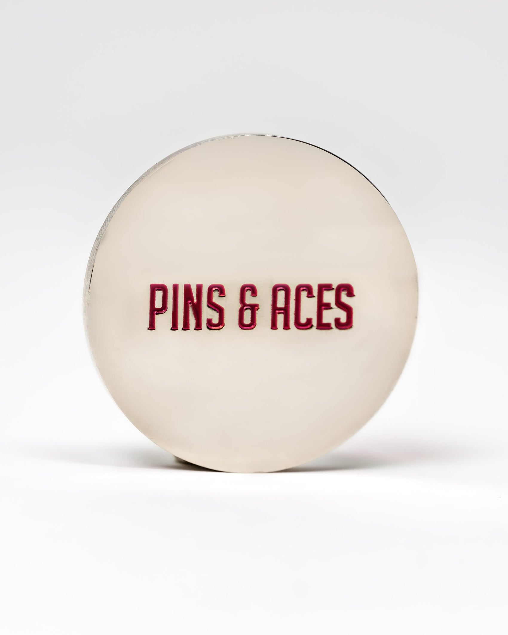 The 6 Milly - Smooth Ball Marker by Pins and Aces is a round, metallic marker with bold red "PINS & ACES" lettering on the front, finished with enamel paint and displayed on a plain white background.