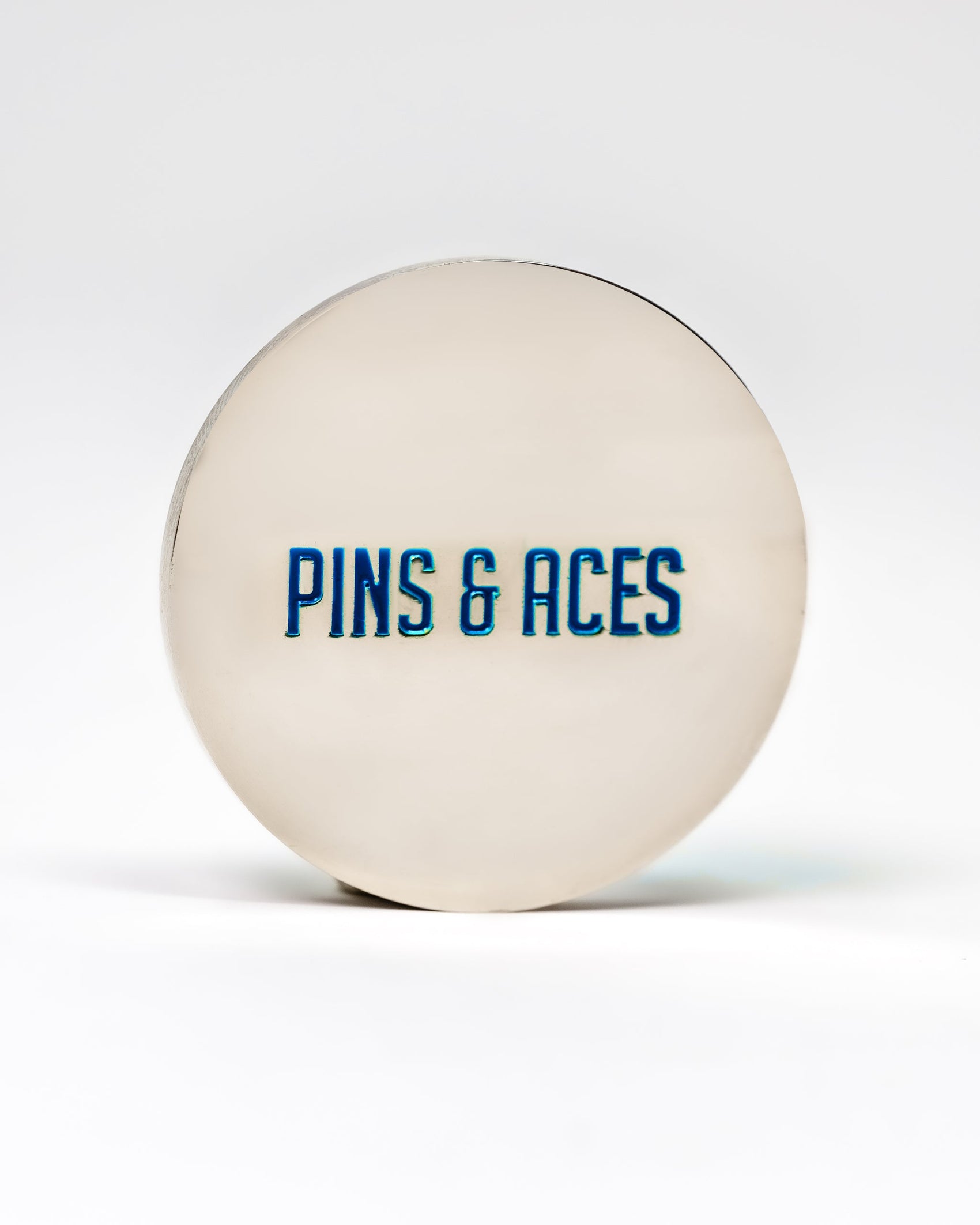 The 6 Milly - Cool Mint Ball Marker by Pins and Aces is a round, silver-colored accessory with the brand's name in blue enamel on the front, designed for marking golf balls, shown on a white background.