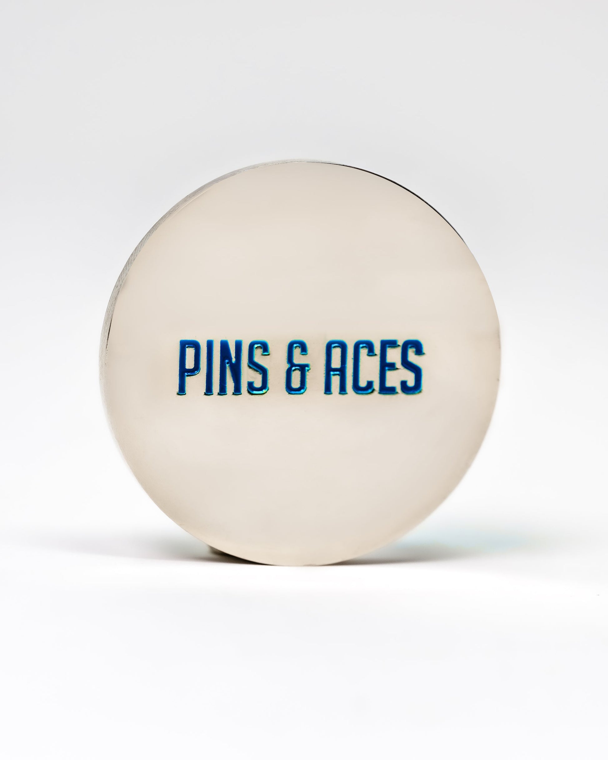 The 6 Milly - Cool Mint Ball Marker by Pins and Aces is a round, silver-colored accessory with the brand's name in blue enamel on the front, designed for marking golf balls, shown on a white background.