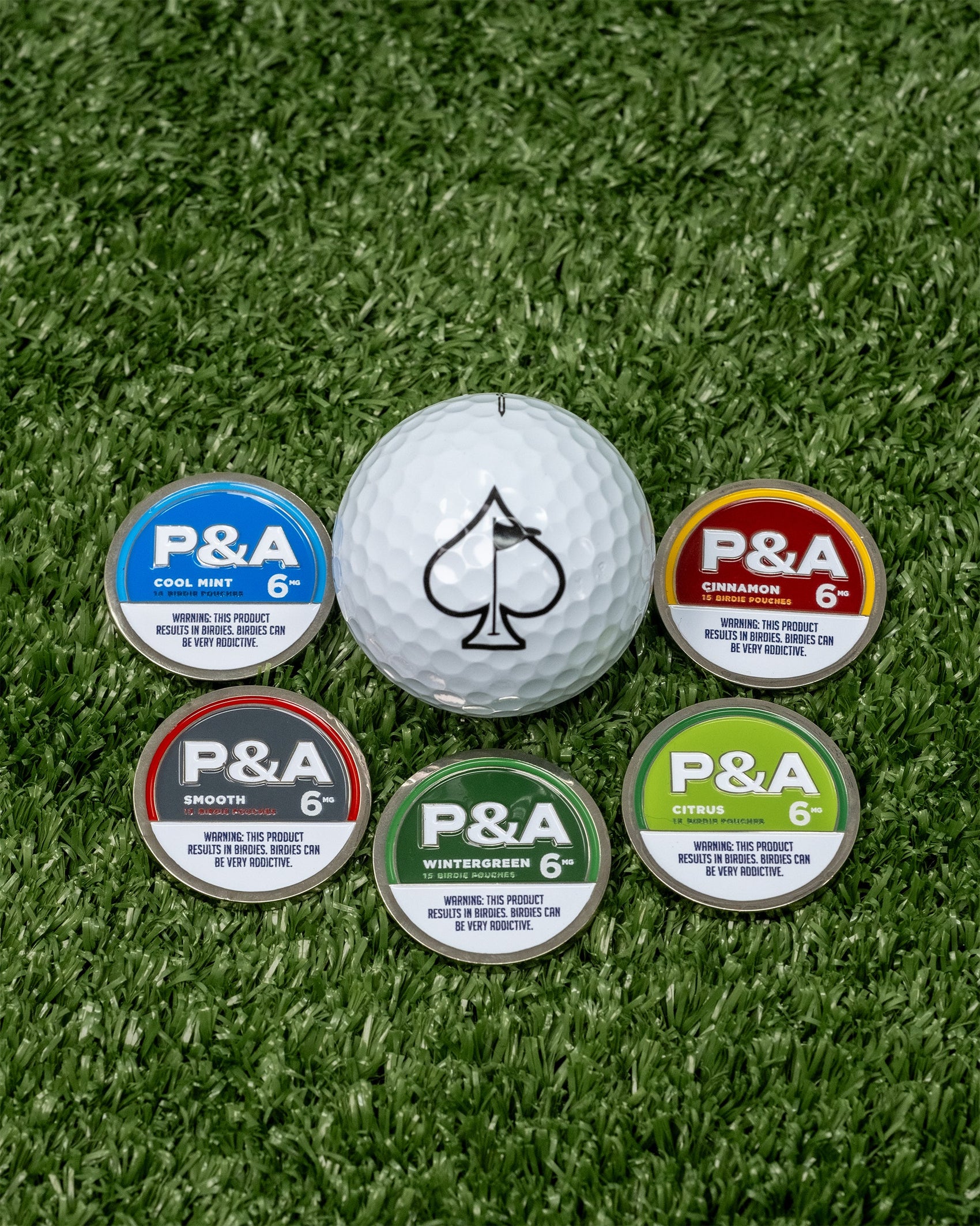 The Pins and Aces 6 Milly - Cinnamon Ball Marker, shaped like a golf ball with a spade symbol, is displayed alongside five tins of P&A nicotine pouches in assorted flavors on green artificial grass.