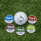 A white Pins and Aces 6 Milly - Wintergreen Ball Marker sits among five P&A tins in Cool Mint, Smooth, Wintergreen, Citrus, and Cinnamon flavors on green turf—ideal for pairing with your favorite golf accessories.