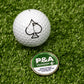 A white golf ball with a black spade symbol sits on artificial grass next to a green and white Pins and Aces 6 Milly - Wintergreen Ball Marker.