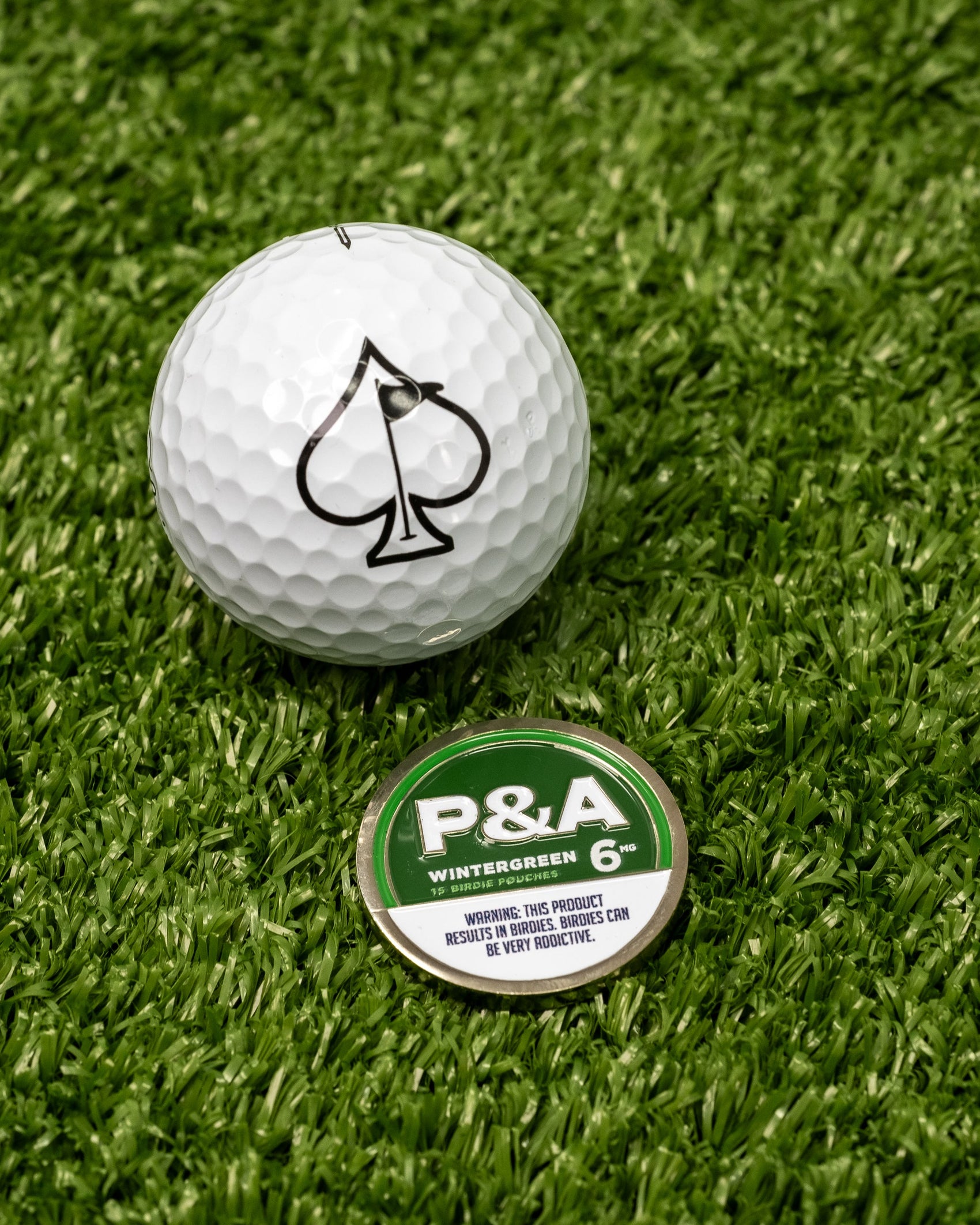 A white golf ball with a black spade symbol sits on artificial grass next to a green and white Pins and Aces 6 Milly - Wintergreen Ball Marker.
