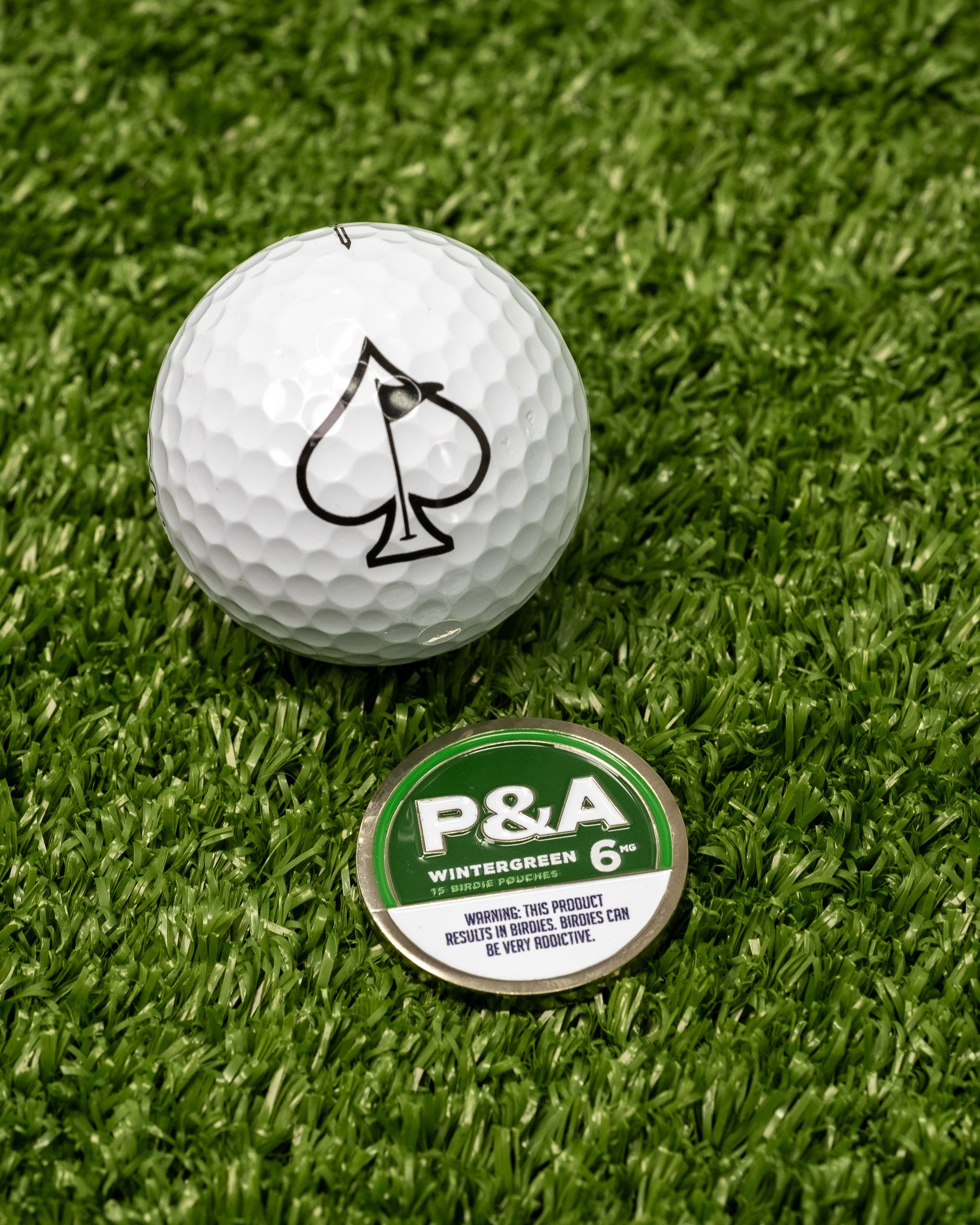 A white golf ball with a black spade symbol sits on artificial grass next to a green and white Pins and Aces 6 Milly - Wintergreen Ball Marker.