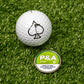 A Pins and Aces 6 Milly - Citrus Ball Marker, with a spade logo golf ball, sits on artificial grass beside its round container featuring a nicotine warning and enamel paint markers on the magnetic lid.