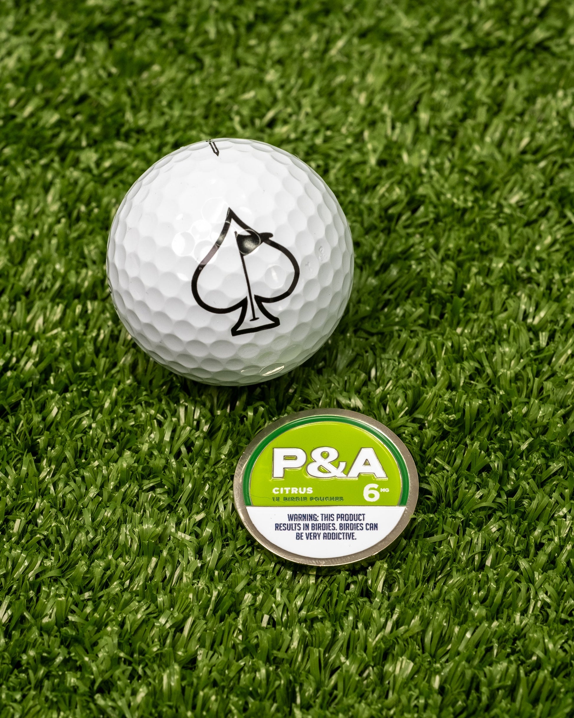 A Pins and Aces 6 Milly - Citrus Ball Marker, with a spade logo golf ball, sits on artificial grass beside its round container featuring a nicotine warning and enamel paint markers on the magnetic lid.