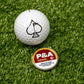 A Pins and Aces 6 Milly - Cinnamon Ball Marker, featuring a spade symbol, sits on green turf beside a tin labeled P&A Cinnamon 6 and an enamel golf marker.