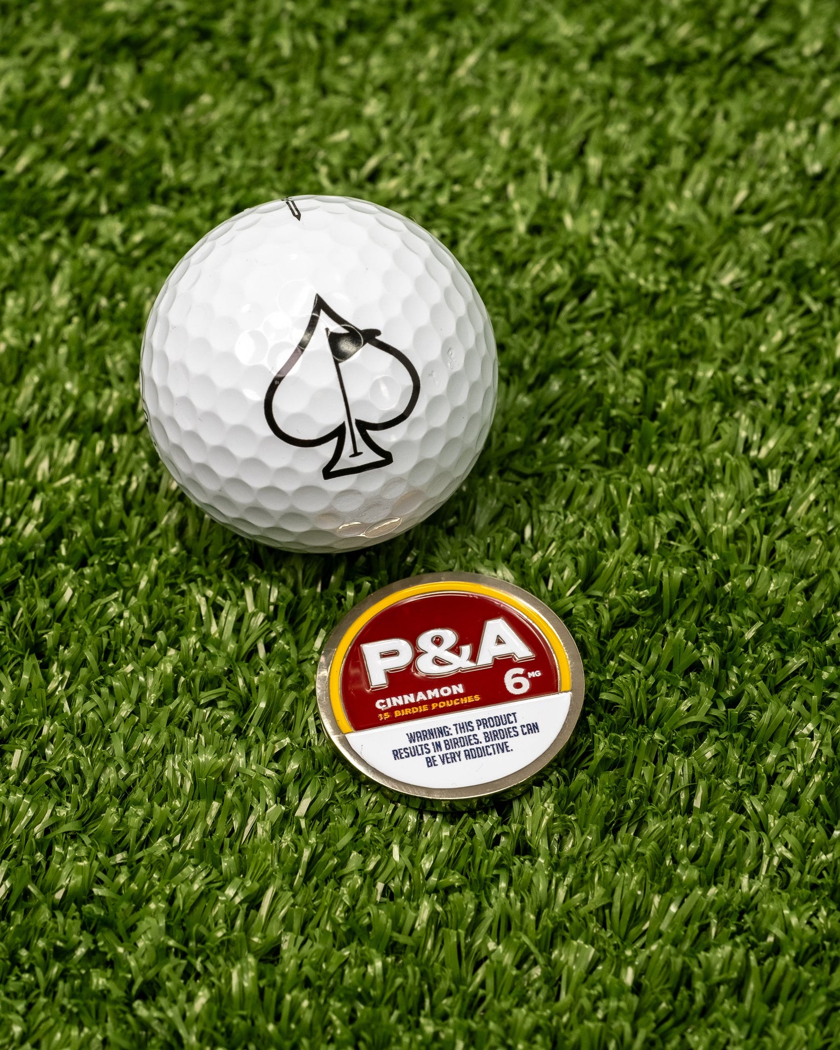 A Pins and Aces 6 Milly - Cinnamon Ball Marker, featuring a spade symbol, sits on green turf beside a tin labeled P&A Cinnamon 6 and an enamel golf marker.
