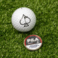 A white golf ball with a black spade sits on turf beside Pins and Aces enamel ball markers, including the 6 Milly - Smooth Ball Marker marked "P&A Smooth 6" with a tobacco/nicotine warning. Ideal for any magnetic surface.