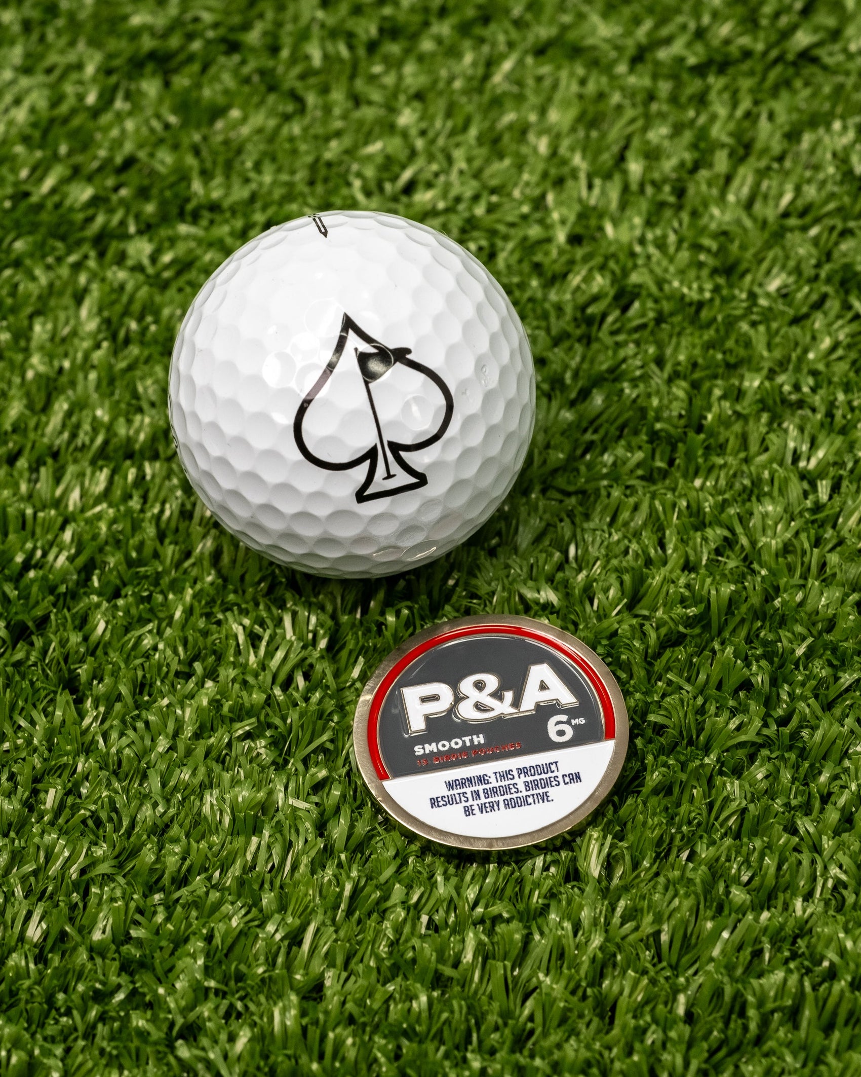A white golf ball with a black spade sits on turf beside Pins and Aces enamel ball markers, including the 6 Milly - Smooth Ball Marker marked "P&A Smooth 6" with a tobacco/nicotine warning. Ideal for any magnetic surface.