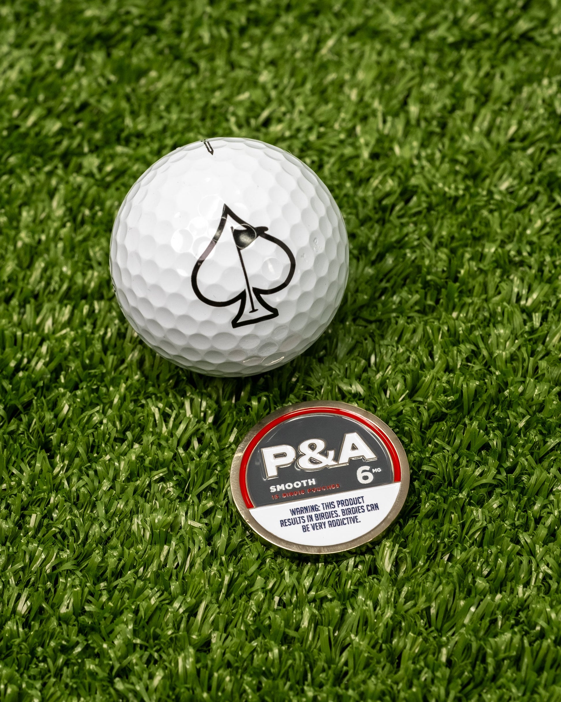 A white golf ball with a black spade sits on turf beside Pins and Aces enamel ball markers, including the 6 Milly - Smooth Ball Marker marked "P&A Smooth 6" with a tobacco/nicotine warning. Ideal for any magnetic surface.