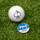A Pins and Aces 6 Milly - Cool Mint Ball Marker in a round blue tin sits on green turf beside a white golf ball with a black spade symbol. The enamel-painted marker adds style to your game.
